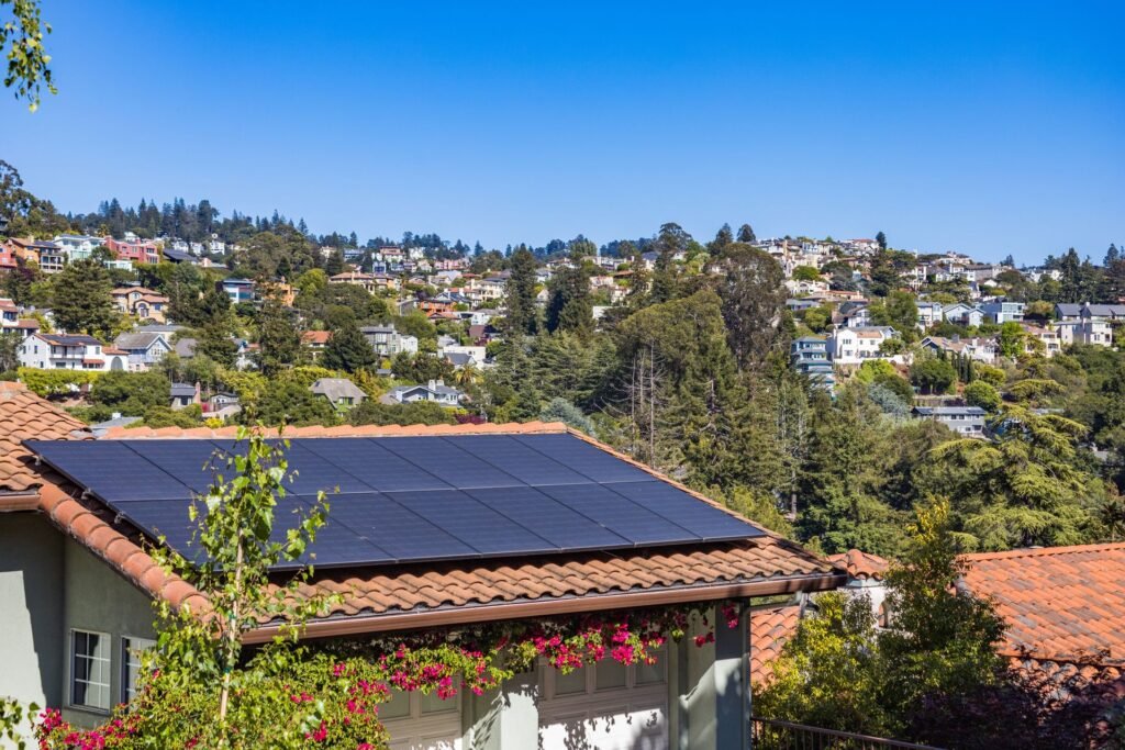 Modulos fotovoltaicos instalados sobre telhado de telhas ceramicas de casa em bairro residencial de encosta, com casas e vegetacao ao fundo sob ceu limpo