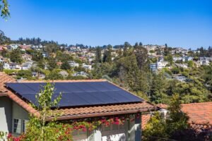 Modulos fotovoltaicos instalados sobre telhado de telhas ceramicas de casa em bairro residencial de encosta, com casas e vegetacao ao fundo sob ceu limpo