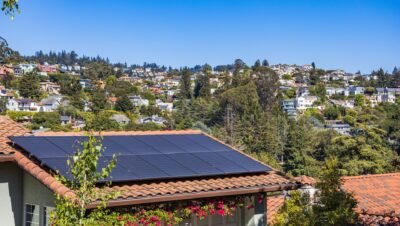 Modulos fotovoltaicos instalados sobre telhado de telhas ceramicas de casa em bairro residencial de encosta, com casas e vegetacao ao fundo sob ceu limpo